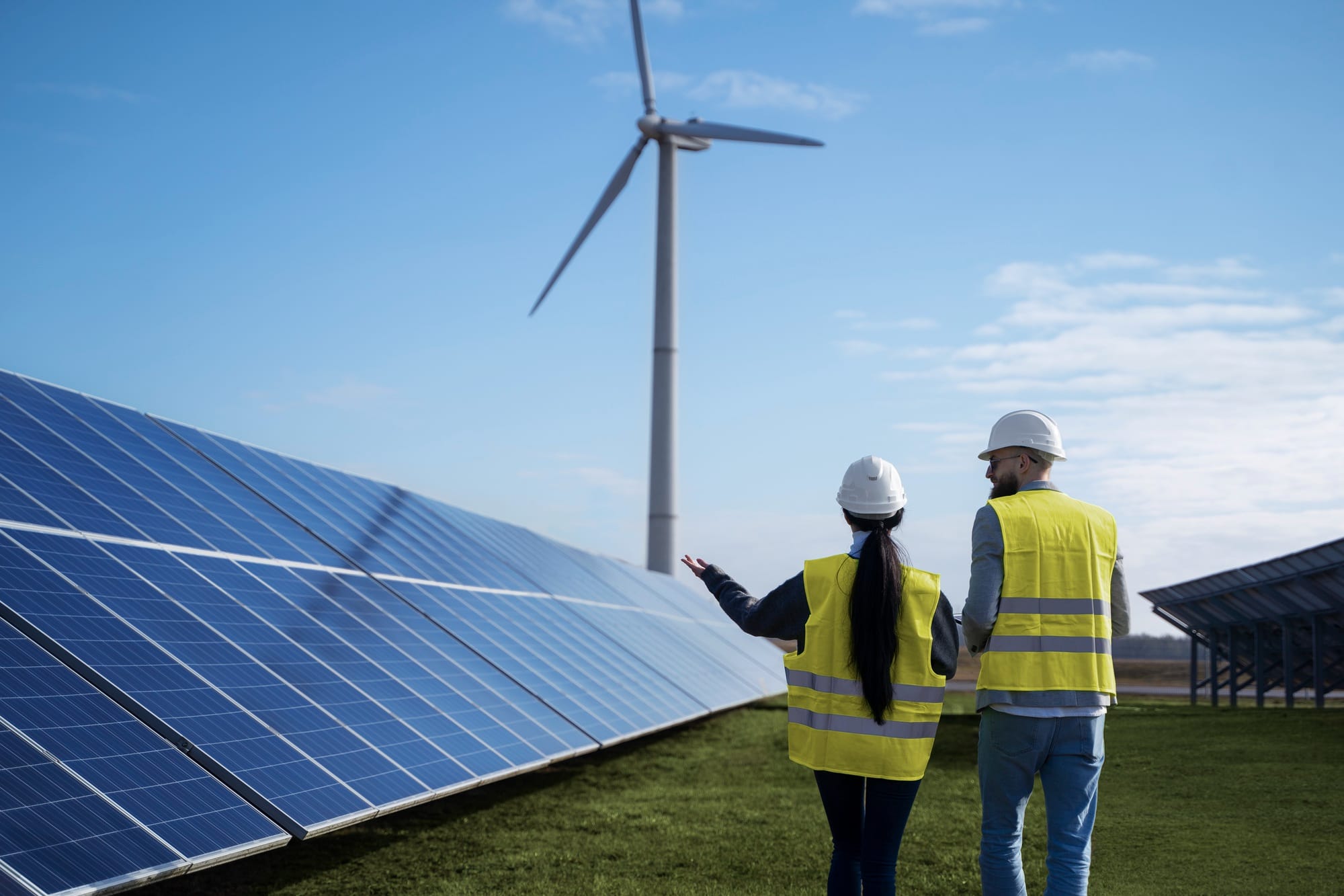 Two workers in yellow vests and hard hats stand near solar panels, with a wind turbine in the background.
